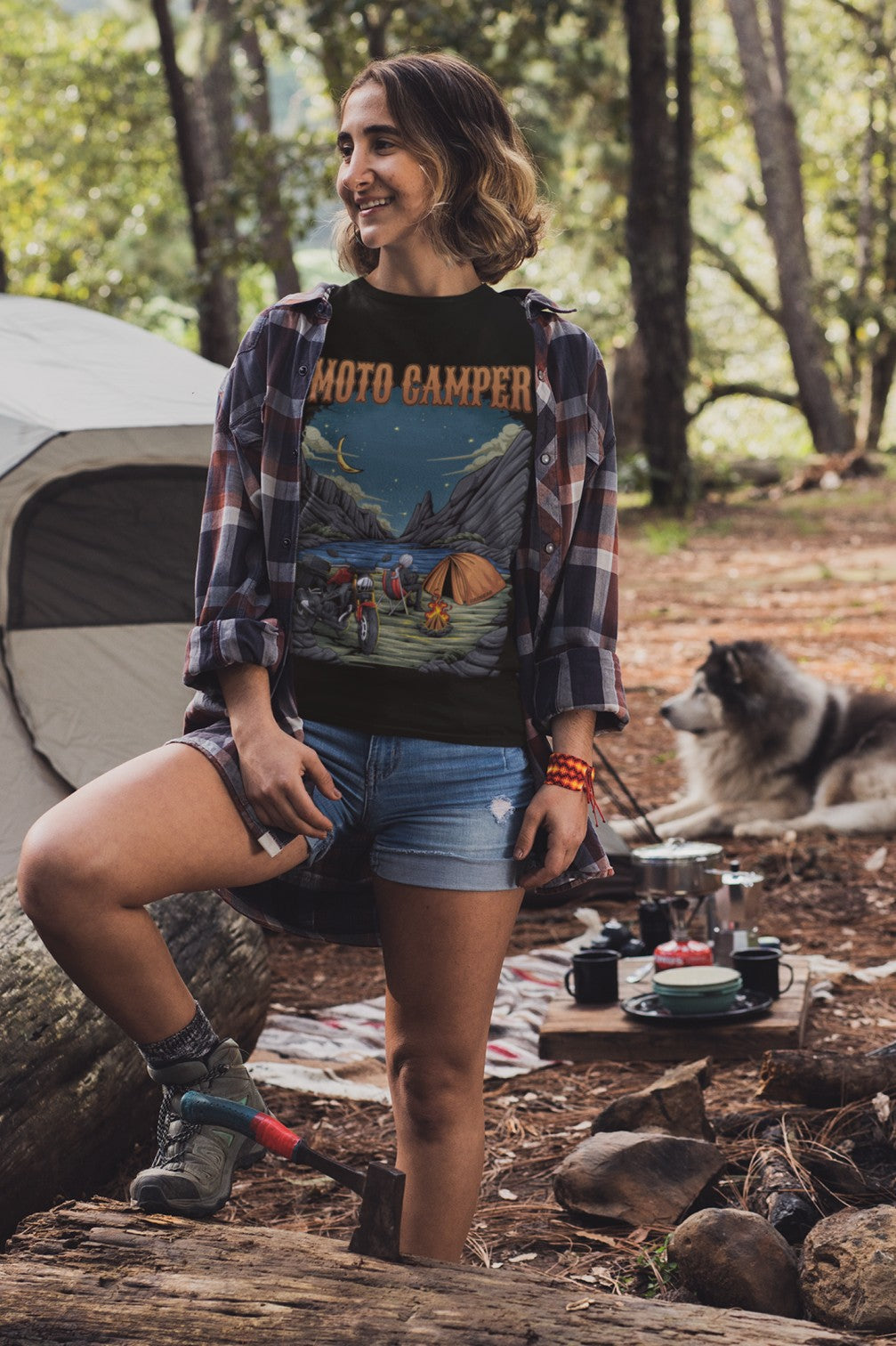 Woman in a plaid shirt and denim shorts standing next to a tent with a dog in a forest setting.